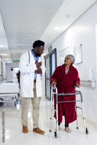 Diverse male doctor and senior female patient with walking frame talking in corridor, copy space