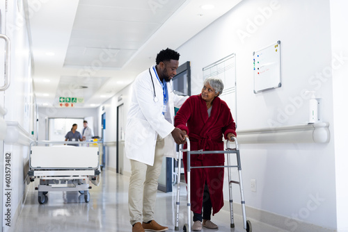 Diverse male doctor helping senior female patient use walking frame in hospital corridor, copy space