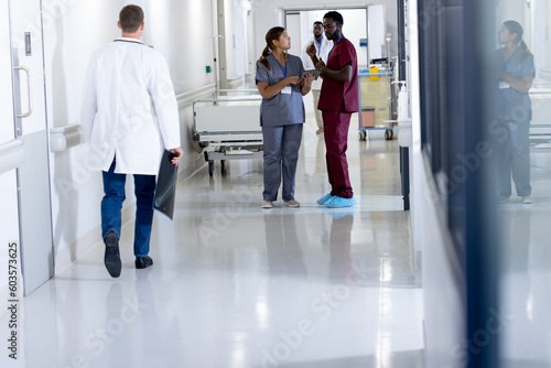 Diverse male and female doctor in scrubs with tablet, talking in busy hospital corridor, copy space