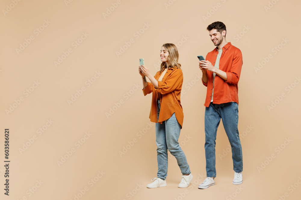 Full body sideways fun young couple two friends family man woman wear casual clothes hold in hand use mobile cell phone together isolated on pastel plain light beige color background studio portrait. Full body sideways fun young couple two friends family man woman wear casual clothes hold in hand use mobile cell phone together isolated on pastel plain light beige color background studio portrait.