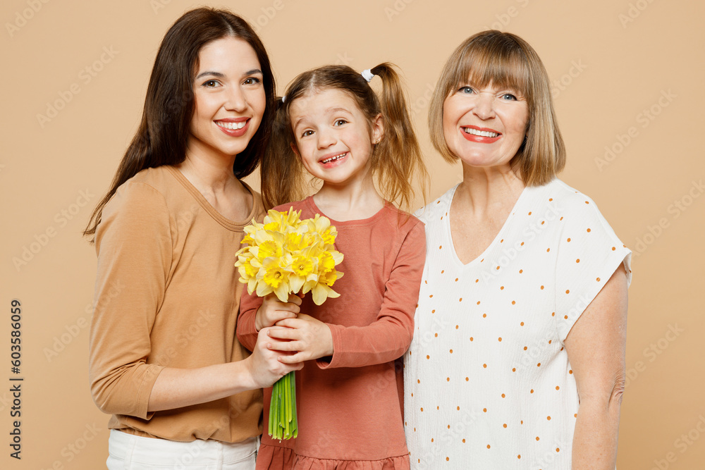 Happy smiling women wear casual clothes with child kid girl 6-7 years old. Granny mother daughter gift giving bouquet of daffodil flowers isolated on plain beige background. Family parent day concept. Happy smiling women wear casual clothes with child kid girl 6-7 years old. Granny mother daughter gift giving bouquet of daffodil flowers isolated on plain beige background. Family parent day concept.