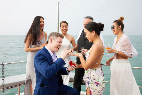 young man showing engagement ring and surprise his girlfriend and friends joined in congratulations or success in a party on luxury yacht