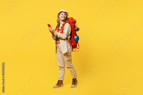 Full body sideways young woman carry bag with stuff mat use mobile cell phone isolated on plain yellow background Tourist lead active lifestyle walk on spare time. Hiking trek rest travel trip concept