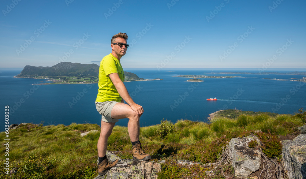 Caucasian tourist with a yellow T-shirt standing on top of Sukkertoppen mountain in Ålesund during summer, enjoying the beautiful view of the surrounding islands and ocean. Norway, day, enjoyment