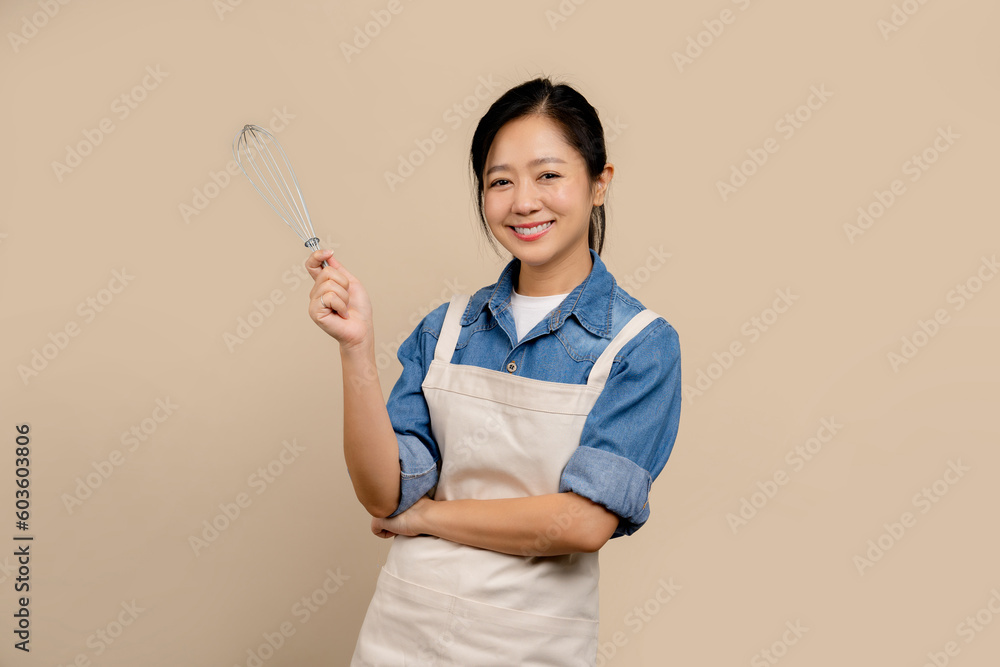 Cheerful Asian woman wearing apron and holding hand whisk standing isolated on light brown background. Cheerful Asian woman wearing apron and holding hand whisk standing isolated on light brown background.