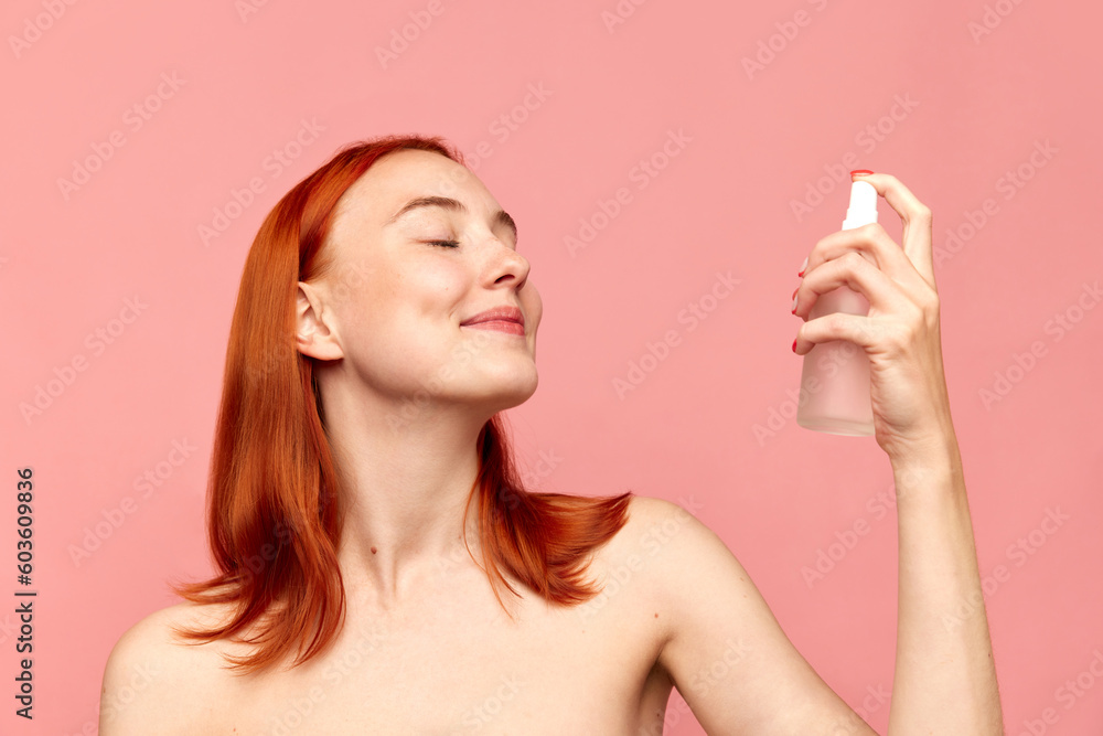 Portrait with redhead girl, model using moisturising spray on face with closed eyes over pink studio background. Facial cosmetics product, masks Portrait with redhead girl, model using moisturising spray on face with closed eyes over pink studio background. Facial cosmetics product, masks