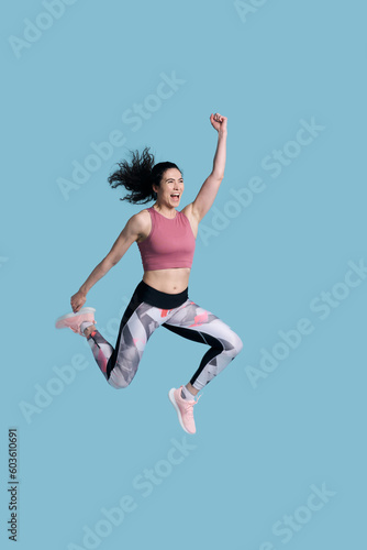 Cheerful determined female athlete in sportswear, smiling and jumping high on blue background