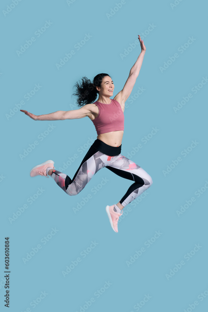 Full size young athletic female fitness trainer doing gym exercises, jumping high on blue background