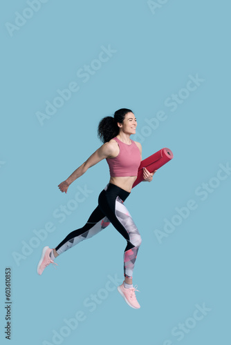 Smiling athletic woman in sportswear carrying yoga or fitness mat while running over blue background