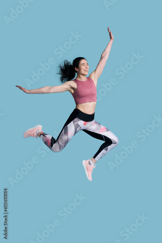 Full size young athletic female fitness trainer doing gym exercises  jumping high on blue background