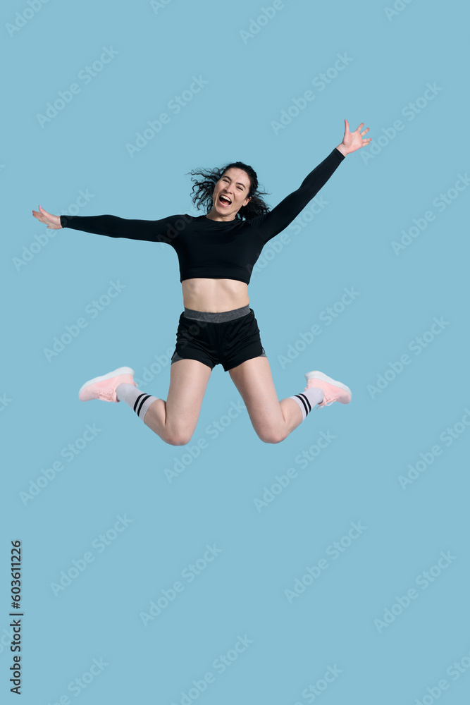 Full length portrait smiling sportive athlete woman, jumping high up over blue isolated background