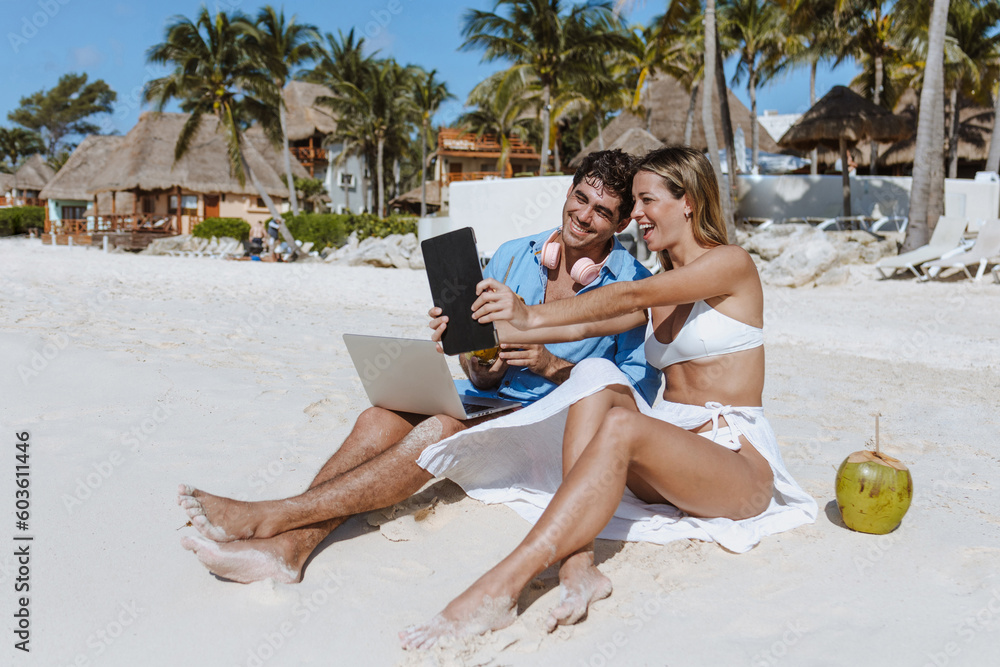 hispanic young couple of digital nomads sit down on the beach with laptop and tablet enjoying the travel adventure vacation and working in Mexico Latin America, Caribbean and Tropical destinations 