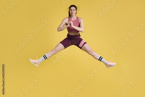 Young sportswoman performing stretching exercises and jumping high up, on isolated yellow background