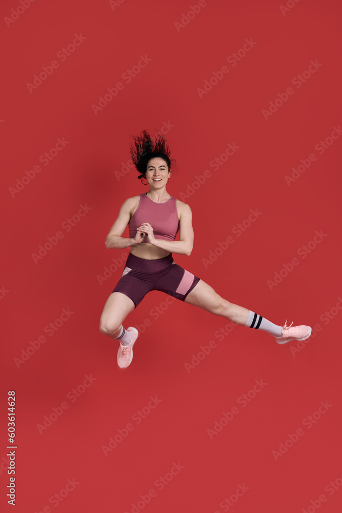 Female athlete in activewear, smiles at camera, performs stretching exercise, jumps over red walll Female athlete in activewear, smiles at camera, performs stretching exercise, jumps over red walll