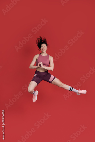 Fototapeta Naklejka Na Ścianę i Meble -  Female athlete in activewear, smiles at camera, performs stretching exercise, jumps over red walll
