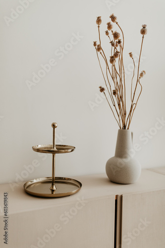 A golden plated cake stand and a beige ceramic vase with dried flowers on the cabinet. Copy space, white background. Modern home decoration.