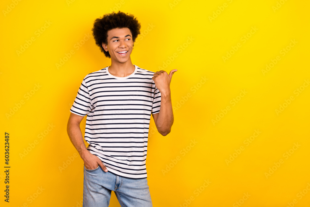 Portrait of cheerful youth guy direct finger empty space wear striped t-shirt denim jeans pants isolated yellow color background Portrait of cheerful youth guy direct finger empty space wear striped t-shirt denim jeans pants isolated yellow color background