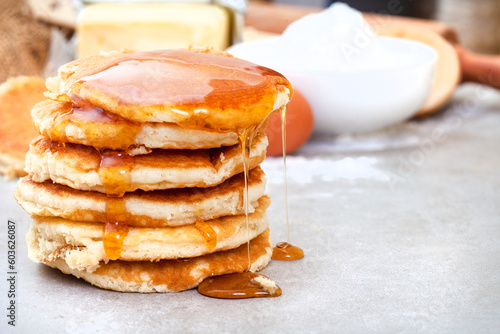 Fresh Pancakes stacked on kitchen table with ingredients in background and with syrup dripping down