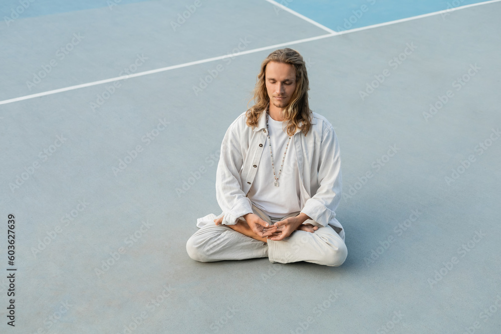long haired man in white cotton clothes practicing yoga in lotus pose on outdoor stadium