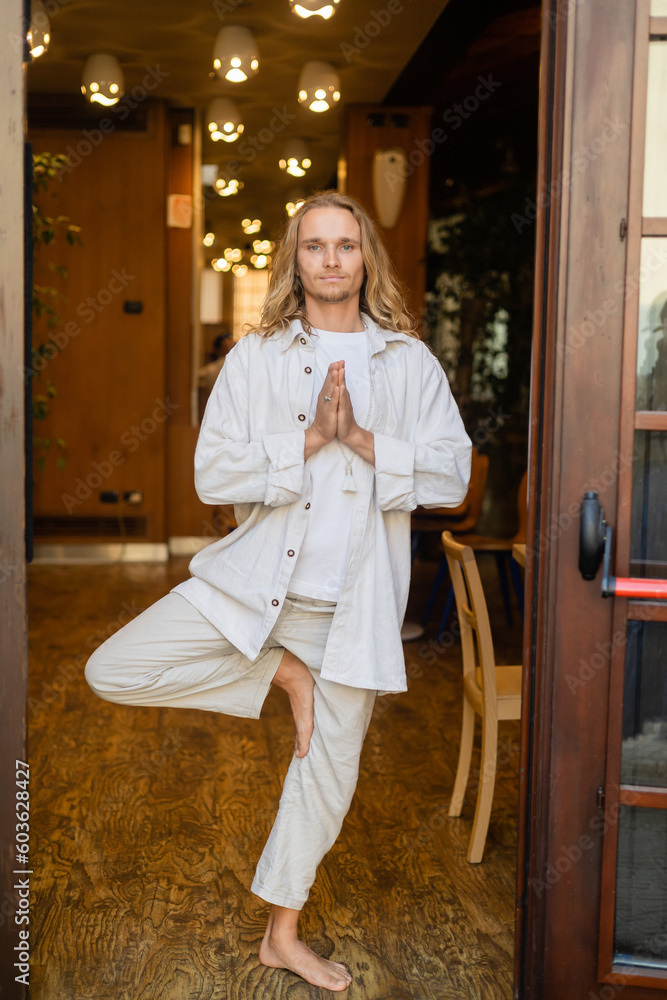 full length of barefoot man in white linen clothes standing in tree pose with praying hands in house with blurred lights