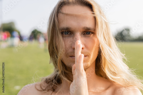portrait of young long haired yoga man holding hand near face and looking at camera outdoors
