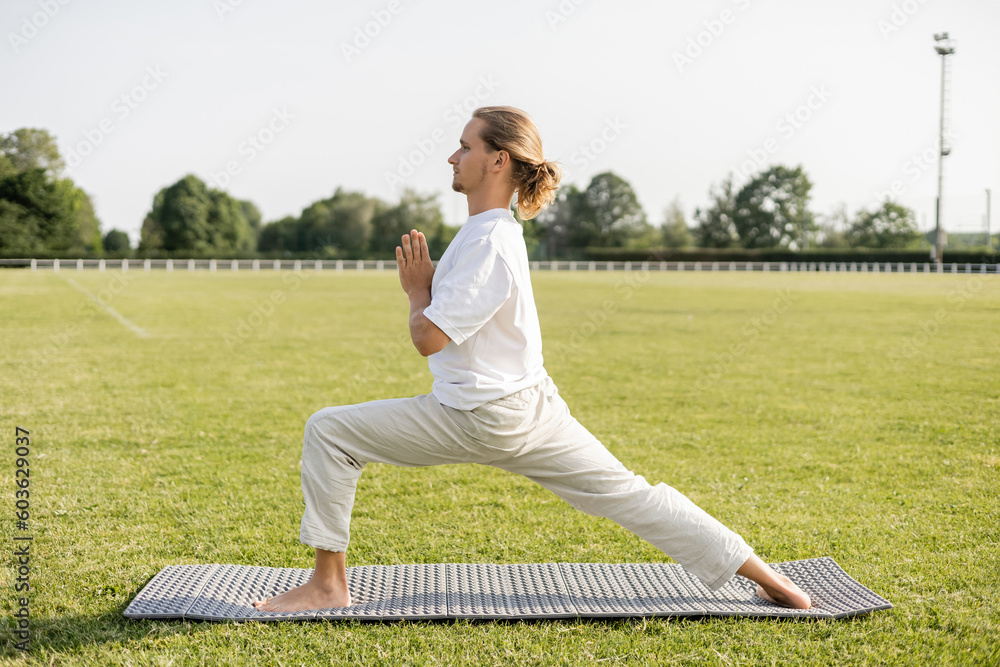 side view of barefoot man practicing yoga in warrior pose with anjali mudra gesture on green grass of outdoor stadium