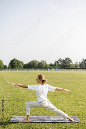 side view of long haired and barefoot man practicing yoga in warrior pose with outstretched hands on grassy field
