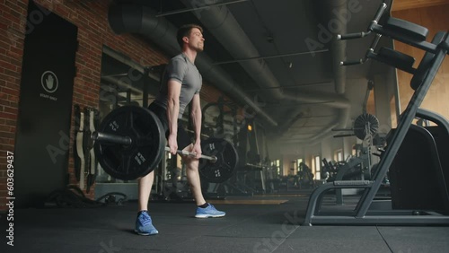 Young handsome athletic man lifts barbell in spacious gym with strength training equipment in black colors. Sportsman performs deadlift, legs far apart. High quality 4k footage