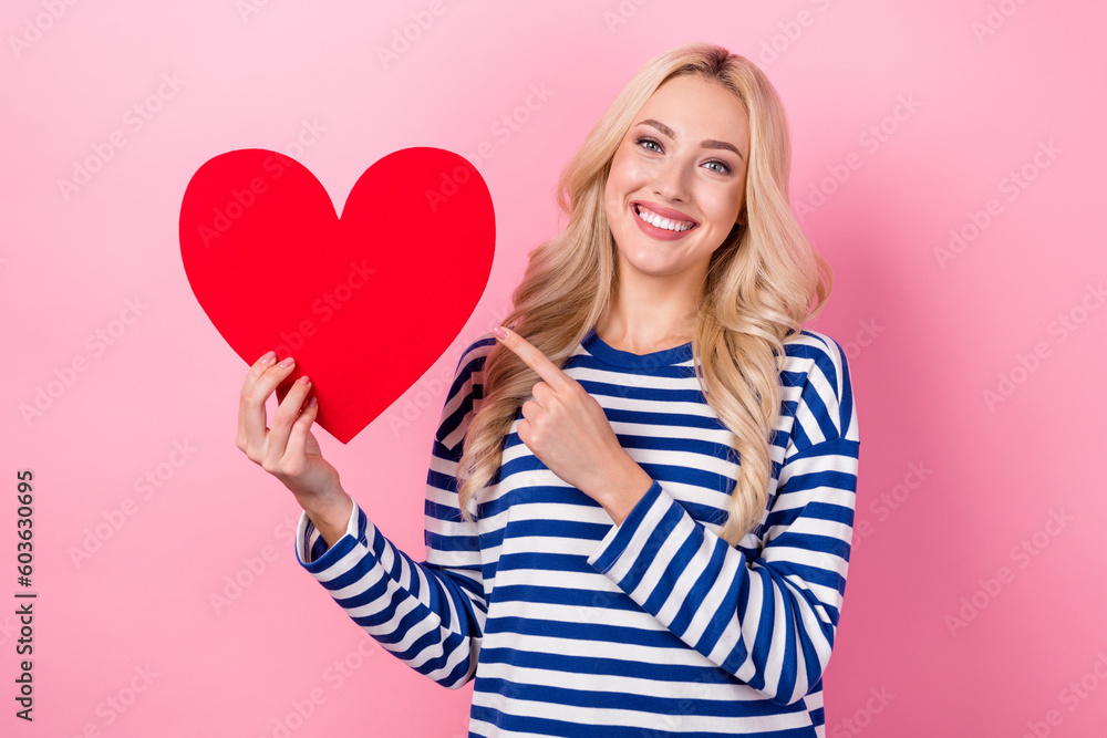 Photo of cheerful positive woman dressed striped shirt pointing red heart empty space isolated pink color background Photo of cheerful positive woman dressed striped shirt pointing red heart empty space isolated pink color background