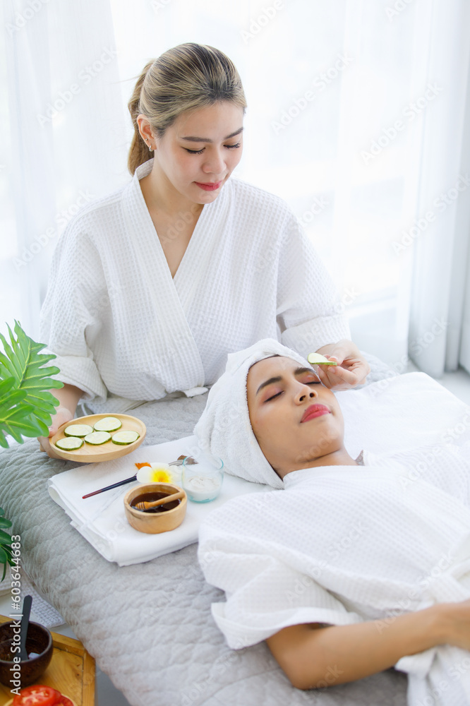 Millennial Asian professional female masseuse therapist in white clean bathrobe standing using sliced cucumber closed customer eyes who laying down on massage bed and massaging face neck and shoulder