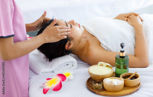 Millennial Asian young beautiful relaxing resting nude naked woman laying lying down on comfortable bed while unrecognizable female masseuse therapist preparing massaging head with oil and hot stones