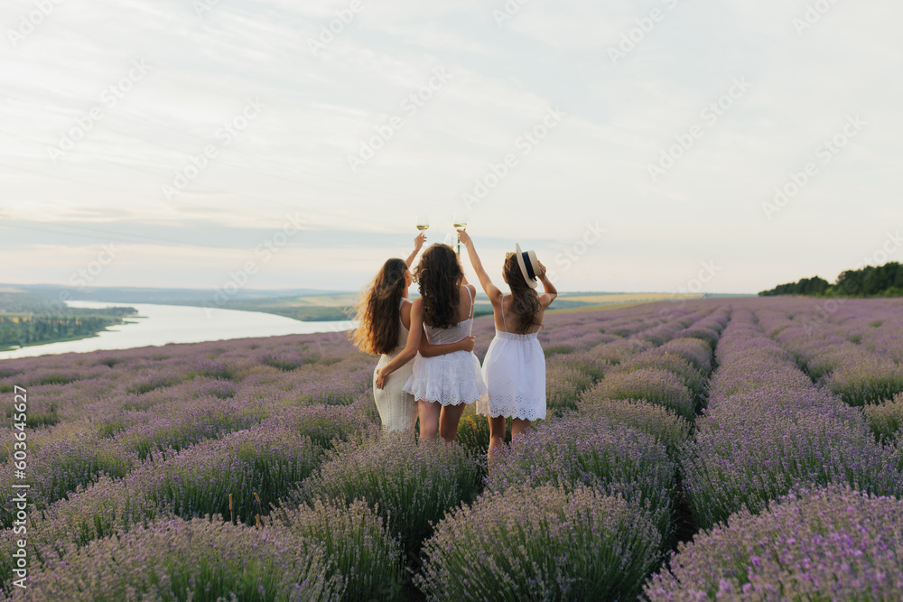Back view of three women in white dresses hold up glasses of wine in a lavender field at summer sunset.