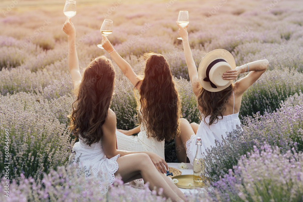Girlfriends having picnic in the lavender field at sunset. Group of young women sitting on lavender field on summer day. Girlfriends drinking wine on outdoor party.