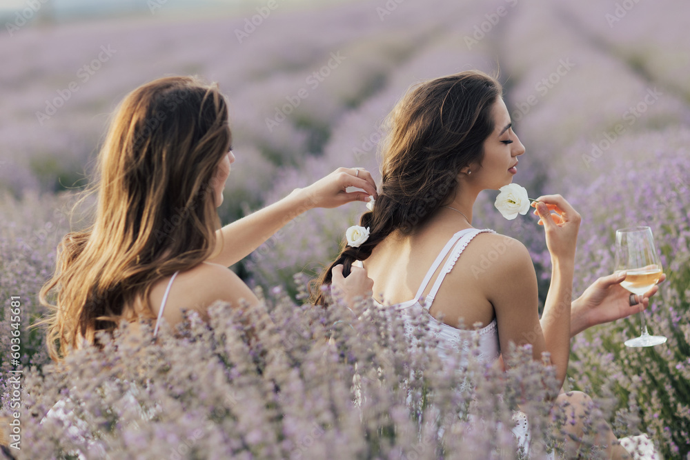 Two sisters enjoying a picnic with fresh fruit and wine while sitting and chatting on field of lavender. One girl braids another girl's hair.