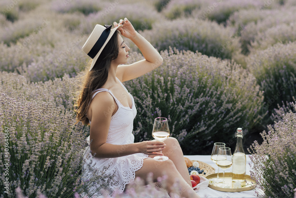 Charming woman in white dress and straw hat holding the glass of wine during the picnic on the lavender field.
