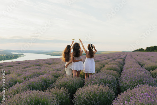 Obraz na płótnie Back view of three women in white dresses hold up glasses of wine in a lavender field at summer sunset. w salonie