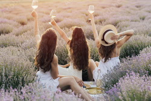 Obraz na płótnie Girlfriends having picnic in the lavender field at sunset. Group of young women sitting on lavender field on summer day. Girlfriends drinking wine on outdoor party.
w salonie