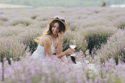 Happy woman sitting in white dress and straw hat sitting in a lavender field with a glass of wine.
