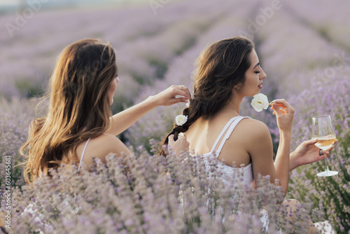 Obraz na płótnie Two sisters enjoying a picnic with fresh fruit and wine while sitting and chatting on field of lavender. One girl braids another girl's hair. w salonie