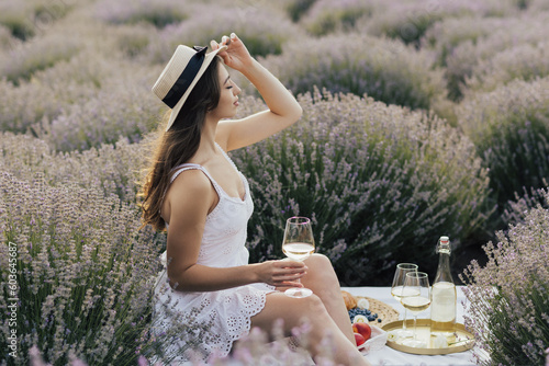 Charming woman in white dress and straw hat holding the glass of wine during the picnic on the lavender field.	
