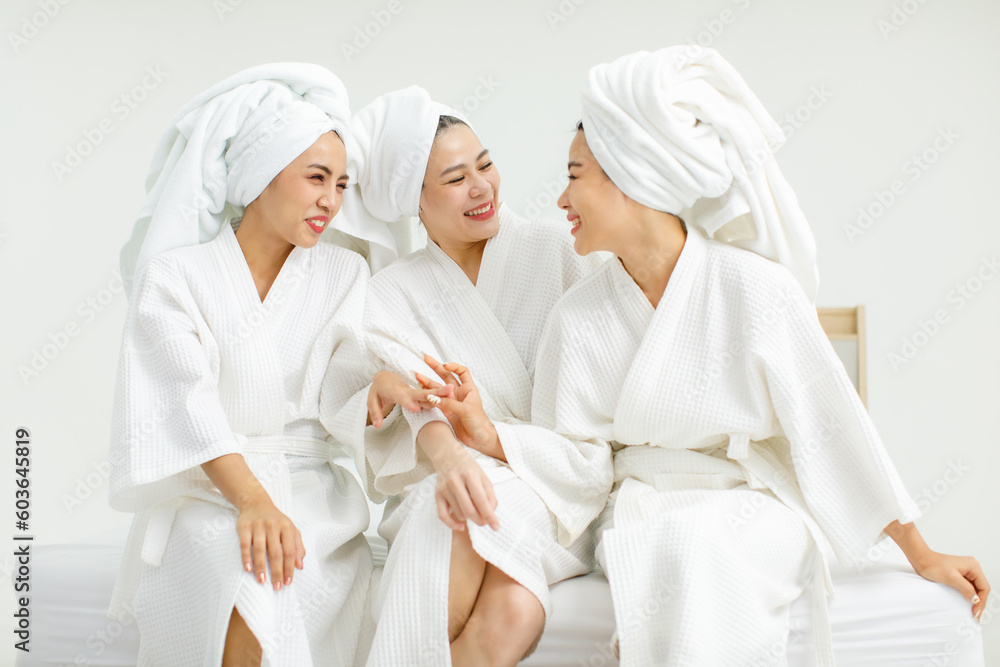 Millennial three Asian female customers friends in white clean bathrobes and towels have appointment at massage resort sitting on chair chit chat talking having conversation together before massaging Millennial three Asian female customers friends in white clean bathrobes and towels have appointment at massage resort sitting on chair chit chat talking having conversation together before massaging