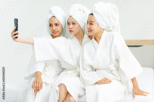 Millennial three Asian female customers friends in white clean bathrobes and towels have appointment at massage resort sitting on chair holding smartphone taking selfie photo together after massaging