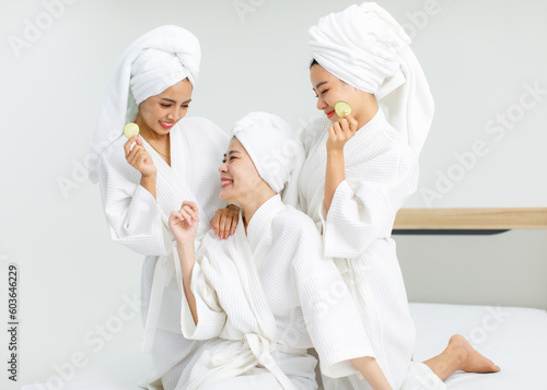 Millennial three Asian female customers friends in white clean bathrobes and towels have appointment at massage resort sit on chair holding sliced cucumbers placed on closed eyes treatment together