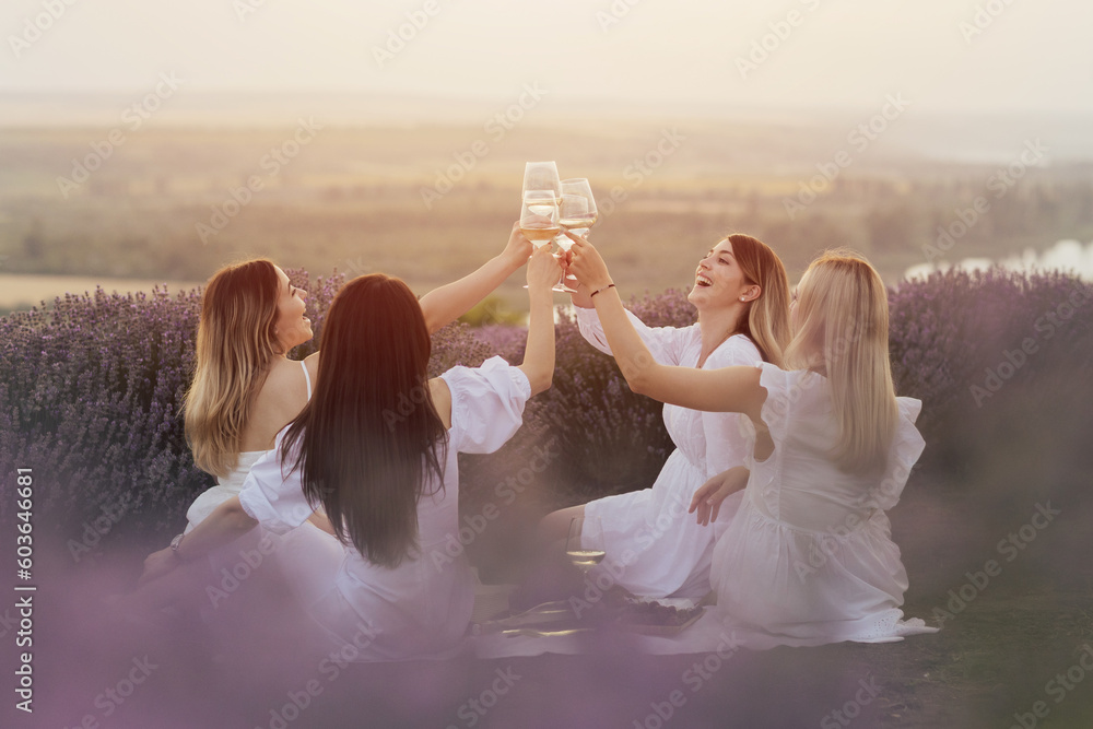 Four women in white dresses are sitting on a blanket and toasting with wine glasses at summer picnic.