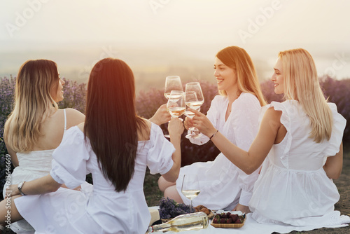 Company of young happy women in white dresses sitting on the picnic in lavender field and clinking wine glasses.