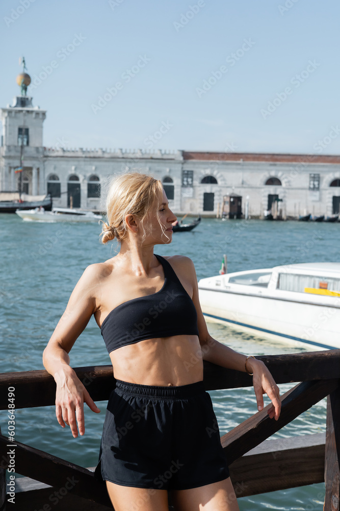 Side view of sportive blonde woman in black crop top and shorts standing on wooden pier in Venice
