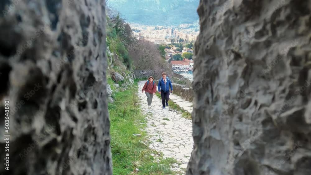 View through ancient fortress wall stone loophole on tourists walk on long cobblestone path TO explore ruins of old town Kotor Montenegro View through ancient fortress wall stone loophole on tourists walk on long cobblestone path TO explore ruins of old town Kotor Montenegro