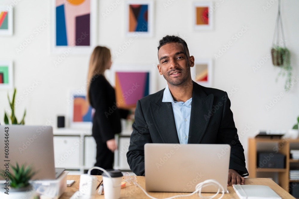 Smiling ethnic businessman in formal clothes sitting at wooden desk with coffee in paper cup and using laptop during workday in light modern office Smiling ethnic businessman in formal clothes sitting at wooden desk with coffee in paper cup and using laptop during workday in light modern office
