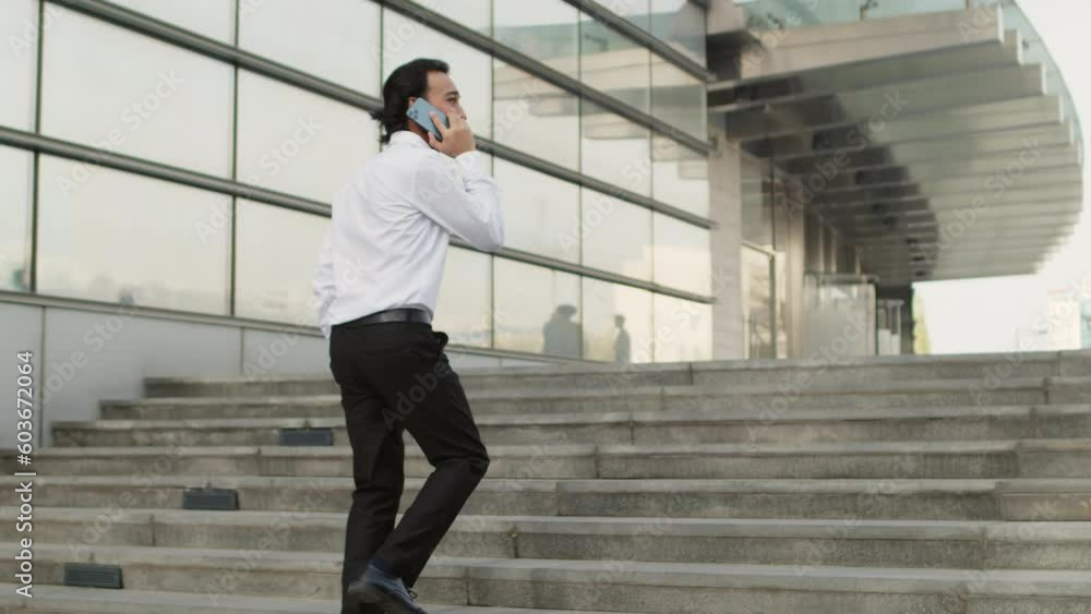Young asian businessman in a white shirt going up the stairs to a business center and solving corporate issues talking on smartphone in a clear day Young asian businessman in a white shirt going up the stairs to a business center and solving corporate issues talking on smartphone in a clear day