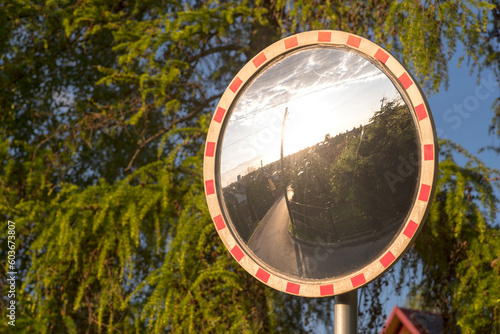 Lustrzany znak drogowy ( u-18) na tle iglastych drzew . W lustrze odbija się wizerunek domów i ulic oraz zachodzące słońce . Znak na peryferiach - Ostrowiec Świetokrzyski - miasta .